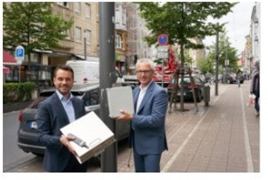 Oberbürgermeister David Langner (links) und evm-Vorstandsvorsitzender Josef Rönz zeigen die Luftsensoren, die an Laternenmasten entlang der Löhrstraße montiert sind, um die Luftqualität zu messen. 